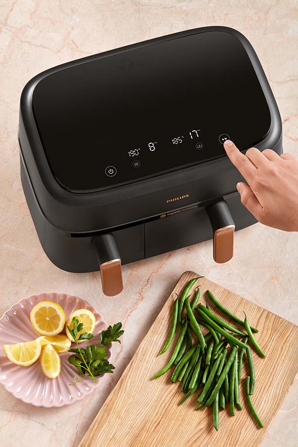 An image of someone using the Philips 3000 Series Air Fryer. It has two different compartments, one of which is larger than the other. There is a hand coming from out of frame to press a button on the air fryer's interface.