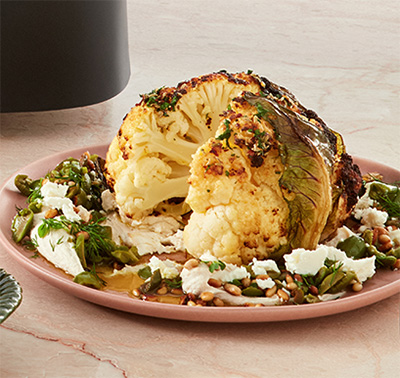 A plate with a whole air-fried cauliflower head on it. The cauliflower has had a wedge taken out of it and has a crispy texture on the outside. It is topped with different herbs and nuts as well as a whipped hummus dressing.