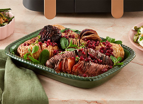 A baking dish full of root vegetables. They have been cut into a hasselback style, meaning they are sliced halfway through to create crispy slices that are still connected at the bottom. The vegetables are topped with pomegranate seeds and herbs.