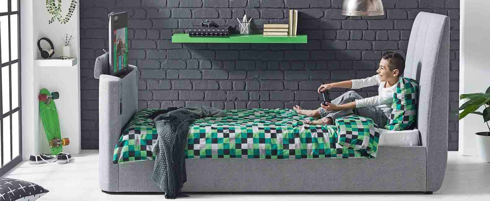 A teen using his bed with hydraulic TV lift to play video games.