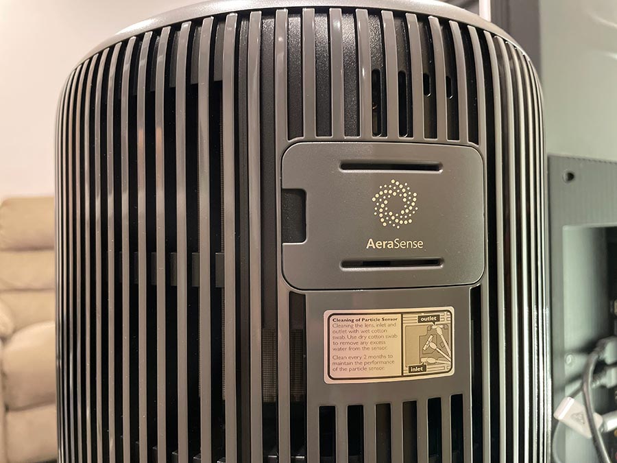 The rear side of an air purifier, mounted in a living room, surrounded by a beige couch and a television.