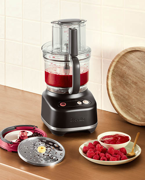 A food processor on a bench in a kitchen. Next to the food processor is a stack of two attachments as well as a plate of fresh raspberries with a bowl of raspberry puree. The food processor also has raspberry puree inside.