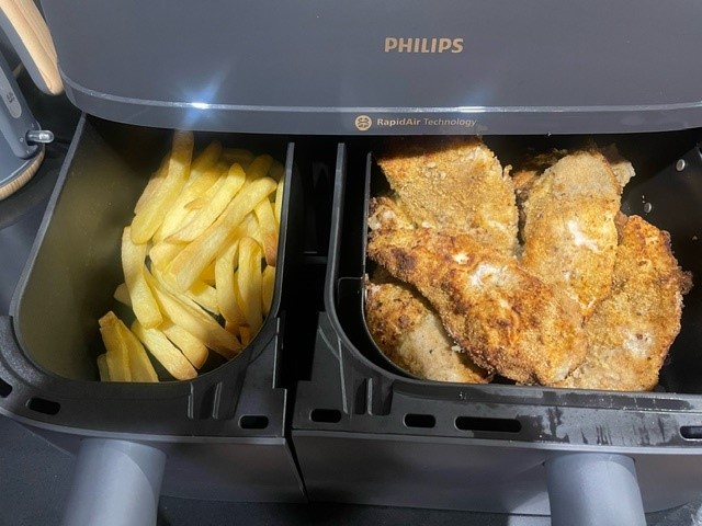 A photo showing the Philips 3000 Series XXXL Air Fryer with its basket open, showing golden potato chips (or 'fries') on the left side and crumbed chicken pieces on the right.